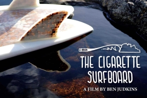 The tail of a surfboard made out of cigarette butts floats on water, the text "The Cigarette Surfboard: A Film by Ben Judkins" is written in the bottom right corner in white