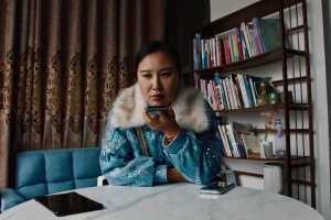 A Chinese woman with dark hair and a sequined blue jacket with a fur collar sits at a white marble table and speaks into her cell phone. Another cell phone rests on the table by her elbow. There is a brown curtain and wooden bookshelf behind her.