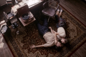 Two people lying on the floor in an office with dark brown hard wood floors, dark brown wooden chairs. One person is wearing long dark pants with a tan short sleeve button up shirt. The other person is wearing a dark plaid skirt with a white shirt sleeve top, ankles socks, with dark brown shoes.
