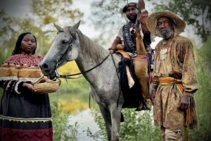 Three Black Seminoles, a woman and two men, wearing traditional clothing. One of the men is sitting on a gray horse while the others stand next to the horse.