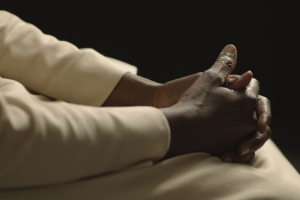 This picture shows a close-up of dark-skinned hands belonging to a Catholic nun dressed in a white robe against a black background. Her face can not be seen. The area in which the shot was taken is an interior illuminated with artificial light creating a high contrast.