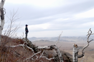In the distance, a young man stands on the edge of a mountain and looks across a wintery valley.