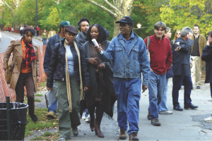 Greaves wearing a black hat with a blue jean jacket and blue jeans, with cast and crew in Central Park during shooting of Symbiopsychotaxiplasm: Take 2 1⁄2 (2003).