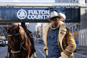 A man wearing a wide brim hat and a shearling jacket stands next to a brown horse in front of the Fulton County Courthouse