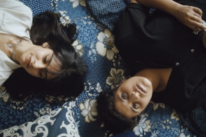 Two women lay on a bed with blue floral bedsheets.