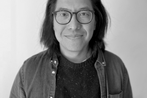 Black and White image of an Asian man. He is wearing black glasses and smiles at the camera. He has shoulder-length black hair and is wearing a dark sweater and button-down shirt.