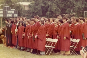 A large group of young men in red robes stand on the football field of their school, preparing for their graduation.