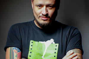 Headshot Rodolfo Castillo Morales with arm tattoos and a black t-shirt with a green and white print