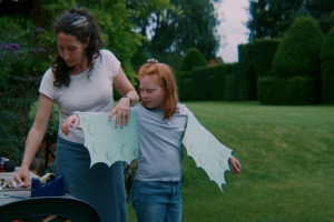 A woman helps a child put on paper wings. They are standing in a grassy field.