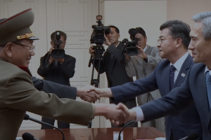 Two officials shake hands as a camera crew films them in the background.