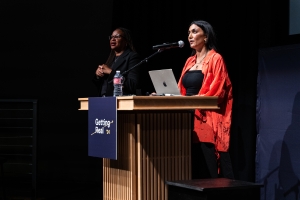Two women stand onstage, one at the podium giving a keynote address.