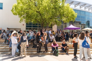 A crowd gathers outside a building. There is a tree with green leaves in the middle of the picture behind the crowd and in front of the building.