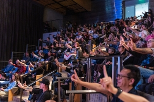 Audience members hold up their pointer finger, looking towards the stage offscreen.