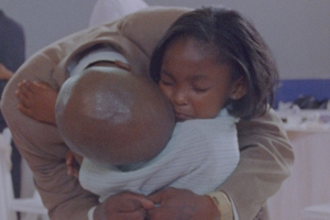 A bald man bends down to hug his daughter. The child is wearing a sad expression.