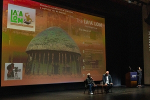 Two men sit at the bottom of a large screen, speaking into microphones.