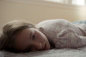 Close up of a teenage girl with light-brown hair and blue eyes, wearing a light-pink sweater and jeans, laying on her stomach on a bedroom floor with a noticeable, hunched-over spine, looking melancholily away from the camera.