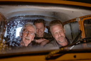 Three white men sit in a car looking out from the windshield 