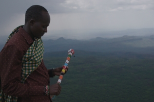 Black man holds a beaded staff and looks over a lush landscape