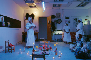 blue light colored a room of three balck women dressed in all white conducting a ritual while a group of people watched 