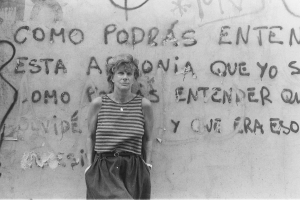 Black and white photo of a woman leaning against a wall. She is a wearing a striped tank top, and her hands are in her pockets. 