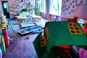 An elementary school classroom in Ukraine lies in ruins following a Russian bombing attack. The windows have been shattered, with glass littering the floor. Shards of glass are scattered across the children's furniture, creating a hazardous environment. In the foreground, a children's playhouse has also been destroyed, highlighting the extent of the devastation.