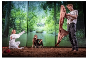 In front of an illuminated screen of the Amazon, three actors sit or stand in the dirt.