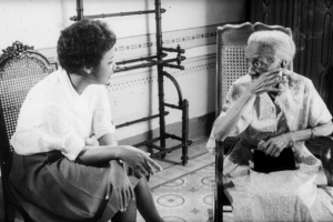 Two Black women sit on whicker chairs, talking cordially to each other.