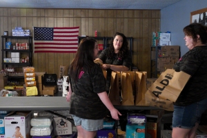 Three individuals carrying items in a pantry, an American flag in the background