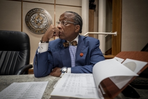 A Black man in a navy suit sits at a wooden courthouse table.