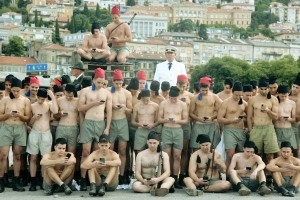 Photograph of a group of young men with bare chests and faded green khaki pants and red caps. A single older man in a white jacket stands in the back row.