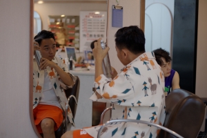 A young Chinese man in a colorful button-up shirt sits in front of a mirror in a barbershop, spraying hairspray onto his head.