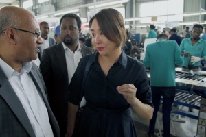 Three people, two men and a woman, are foregrounded speaking to each other. A busy factory assembly line is pictured behind them.