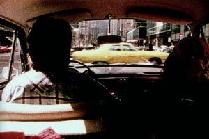 Silhouette of a man driving, with a woman seated on his right. A yellow NYC taxi drives past, visible through the windshield.