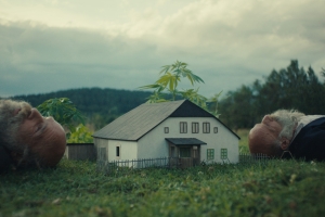 In a verdant landscape, a small house sits postioned between two men lying on their backs and looking up at the sky. 