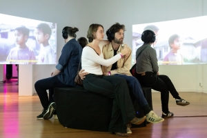 Five people sit on a bench, looking at an installation of videos featuring young Vietnamese children.