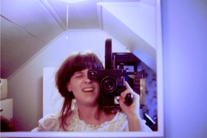 In a mirror, a woman holding a camera on her shoulder is reflected, squinting through the viewfinder and smiling brightly. 