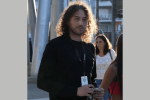 A man with shoulder-length hair stands looking at the camera as the sun illuminates him. 