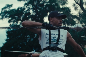 A drum major marches forward with a mace in hand. There are trees behind him.