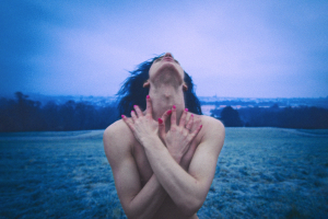 A still from the film shows Adelina standing alone in an open field, her head tilted upward toward the sky. Her bare shoulders and tightly crossed arms convey exposure and restraint, while her upward gaze suggests longing and a deep desire for freedom. The cold blue tones of the landscape and the distant city reinforce her emotional isolation.