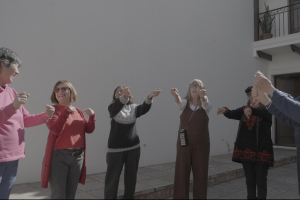Seven women in their 70s and 80s stand in a semi-circle doing stretches in a courtyard bathed in sunlight.