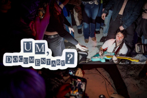 Eli, the frontman of Um, Jennifer?, is on the ground playing a blue guitar. He is surrounded by a swarm of energetic fans recording and cheering him on. The image is captured from an Um, Jennifer? show in Brooklyn. He is wearing gray trousers and a white blouse.