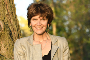 A woman with white skin smiles at the camera. She has short, curly salt-and-pepper hair, blue eyes, red lipstick, and is wearing a quilted black leather blazer. Headshot of Alison McAlpine