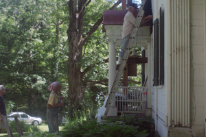 Two female sisters in their 60s and 70s look on as their brother fixes the siding of an old white house on a ladder.