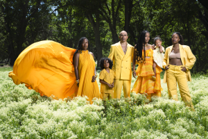 A family of six stands together in a sunlit field of soft white wildflowers, surrounded by lush green grass and tall, dark trees. At the center, a man and woman stand close, with four children gathered around them—one held in the woman’s arms, others standing confidently at their sides. All are dressed in coordinated, flowing and tailored outfits in vibrant shades of yellow and gold, creating a striking contrast against the deep green landscape. One figure on the left wears a dramatic, billowing gown that e
