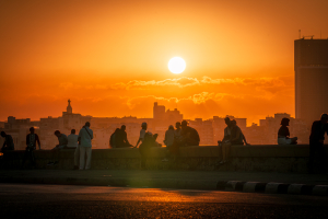 Silhouettes of people sitting and standing along the Malecón seawall in Havana at sunset, with the city skyline behind them and a deep orange sky.