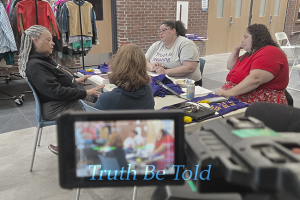 Seen through a camera's viewfinder, four women gather around a table — one wearing a shirt that reads "Truth is Healing" — as Truth Be Told documents stories Vermont has never officially heard before.