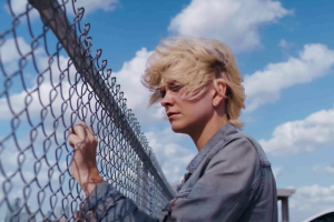  a young person with short, windblown blond hair standing beside a chain-link fence under a bright blue sky with large white clouds. They are wearing a gray denim-style jacket and appear to be looking through or past the fence with a quiet, reflective expression.