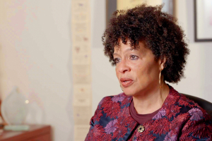 African American woman in her fifties, seated at a desk wearing a top featuring red and pink flower design. Picture frames are visible, but out of focus, in the background.