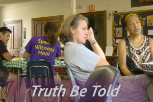 A man on the left in a black shirt at a table with a woman who is wearing a purple shirt with the words on the back: Speak, Witness, Heal.  A woman featured in the center of the photo is leaning back in her chair, legs crossed with her hand on her head looking off camera.  A black woman with a gold hair band is sitting next to the woman looking off camera.