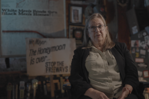 Image shows Kate Kelly, an older woman and resident of the brookland neighborhood who was a witness to the protests at the time. She is sitting being interviewed with a poster and signage from the protests blurred but still visible behind her. They say; "Mr. Mayor the honeymoon is over...stop the freeways!" and "White men's roads through black men's homes!"